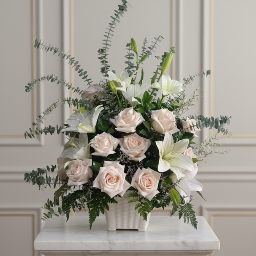 Arreglo de rosas color crema y lirios blancos con follaje verde en maceta blanca sobre mesa de mármol