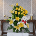 arreglo floral con rosas amarillas, gerberas y lirios blancos acompañado de un oso de peluche blanco con moño rojo sobre pedestal de mármol