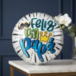 Globo metálico redondo plateado con texto Feliz Día Papá, bigote negro y corona amarilla, apoyado sobre una mesa de mármol con pared azul y orquídea blanca de fondo