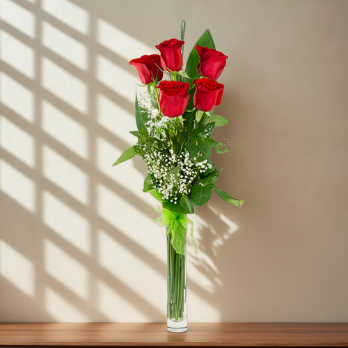 Seis rosas rojas con follaje verde y flores blancas pequeñas en un jarrón de vidrio alto y estrecho sobre una repisa de madera