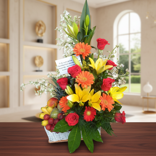 Arreglo floral vertical con lirios amarillos, gerberas naranjas y rosas rojas acompañado de frutas variadas en una canasta blanca sobre mesa de madera, con fondo de sala elegante y ventana arqueada