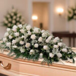 Centro de rosas blancas con follaje verde colocado sobre un ataúd en una sala velatoria
