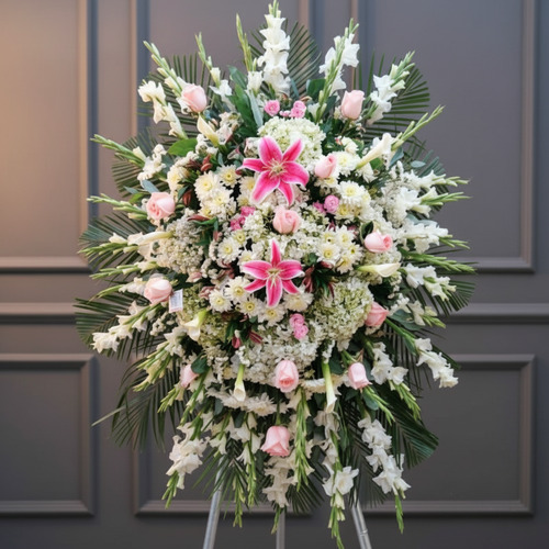 Arreglo floral de pie con lirios rosas al centro, rosas rosadas y flores blancas rodeadas de follaje verde sobre fondo de pared gris