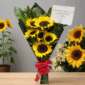 Ramo de girasoles amarillos envueltos en papel negro con lazo rojo y tarjeta de dedicatoria sobre mesa de madera, con macetas de girasoles de fondo
