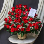 Canasta de mimbre sobre mesa redonda con gran arreglo de rosas rojas y flores blancas de relleno, acompañadas de tarjeta sobre fondo de cortinas blancas y tela gris