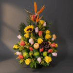 Arreglo floral vertical con rosas de colores, girasol, aves del paraíso y frutas como uvas, manzana y limón sobre fondo gris