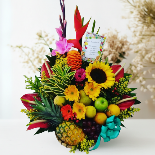 Arreglo floral con frutas tropicales que incluye piña, manzanas, uvas, girasoles y flores de colores en una canasta decorada con lazo turquesa y tarjeta