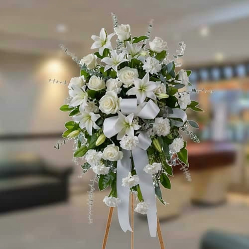 Corona fúnebre de rosas y lirios blancos con lazo blanco sobre caballete de madera en interior